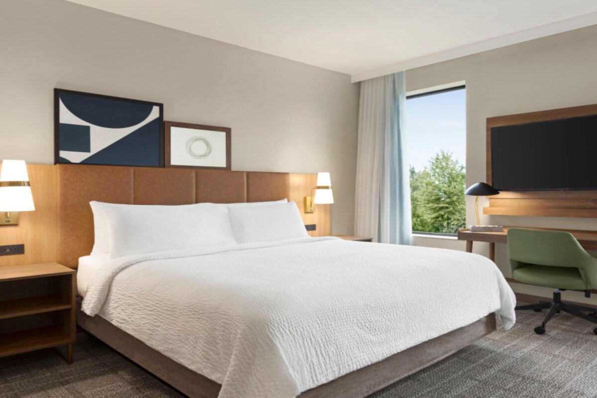 A hotel room with a large bed, two bedside tables with lamps, a desk with a chair, a flat-screen TV, and a window with a view of trees.