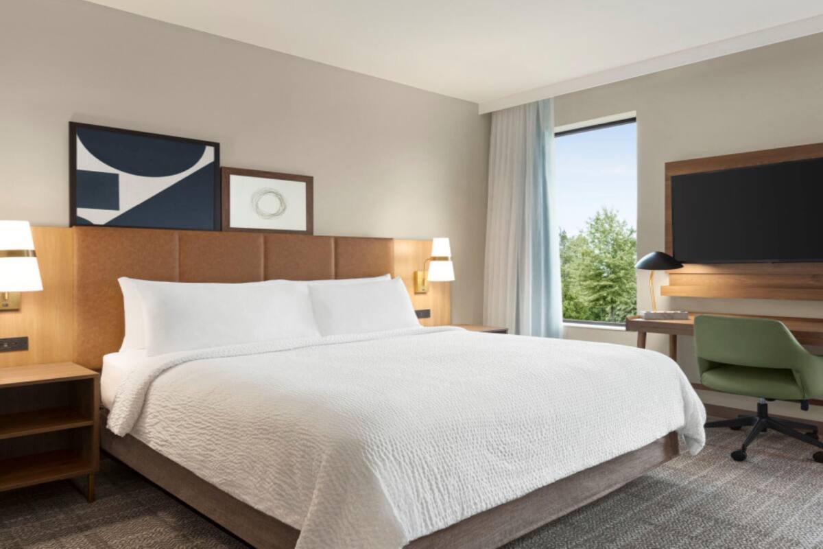 A hotel room with a large bed, two bedside tables with lamps, a desk with a chair, a flat-screen TV, and a window with a view of trees.