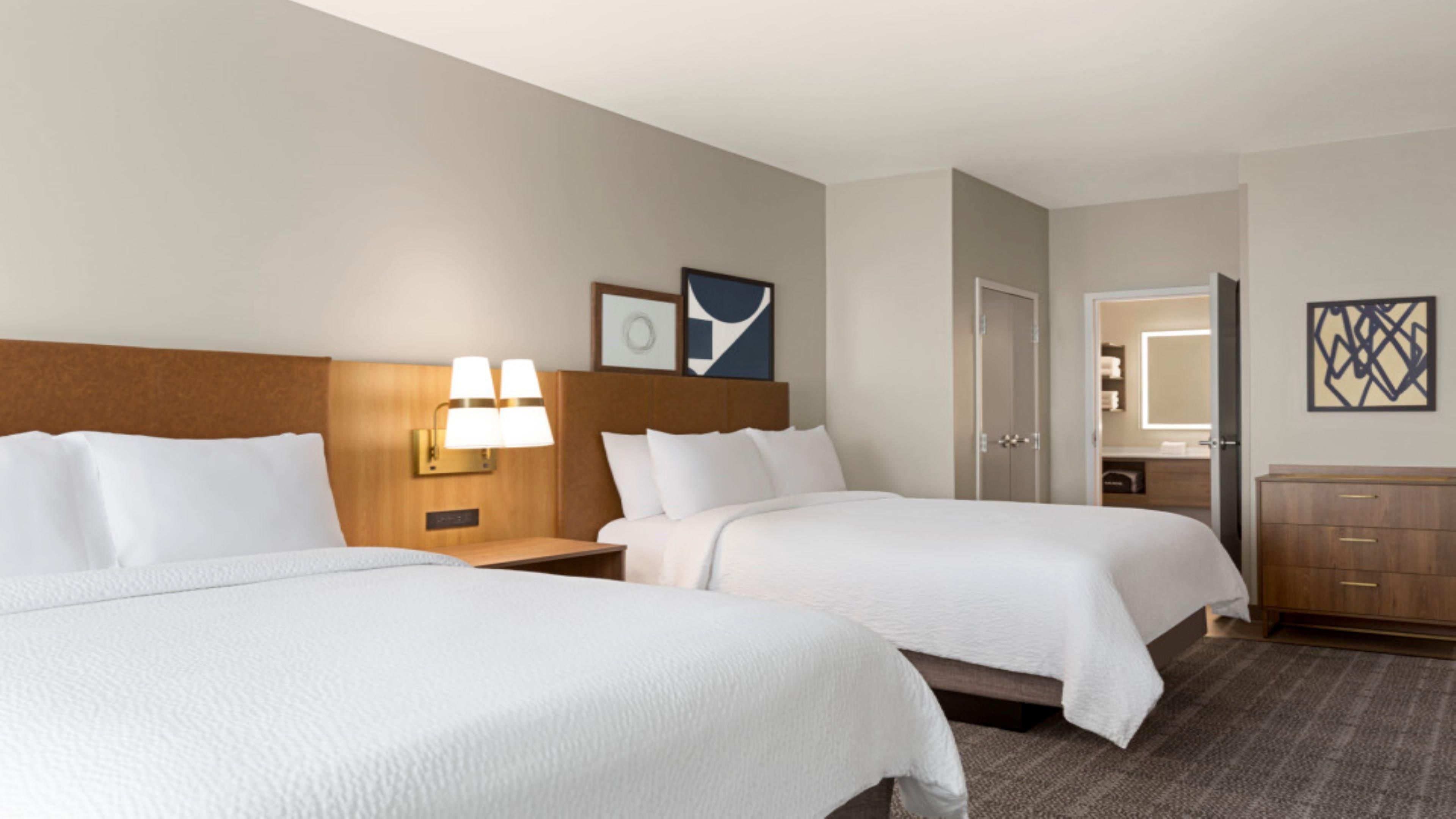 A hotel room with two beds, a nightstand with lamps, a door, a bathroom mirror, and a dresser.