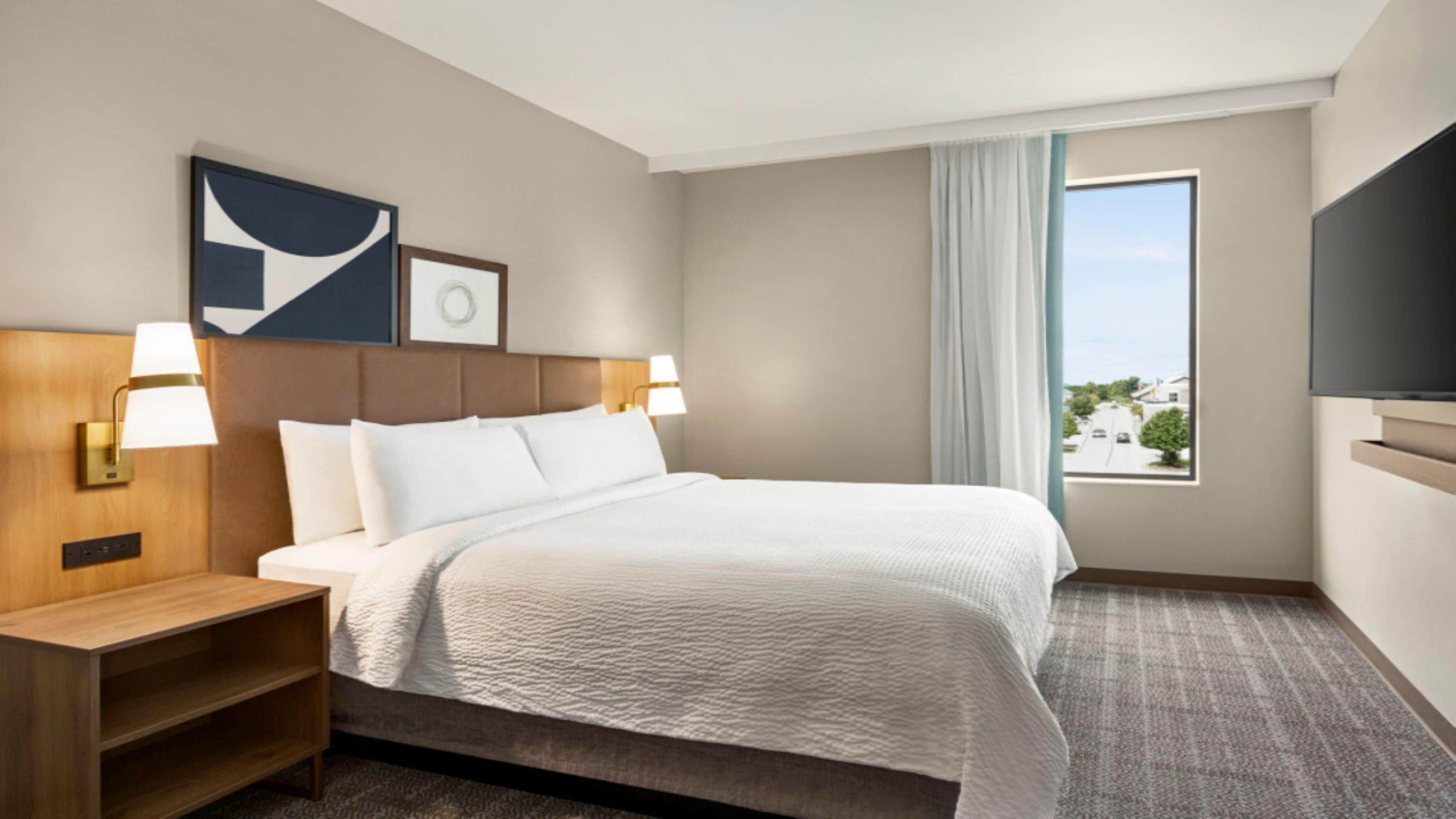 A hotel room with a bed, two bedside lamps, a nightstand, a TV, and a window with a view.