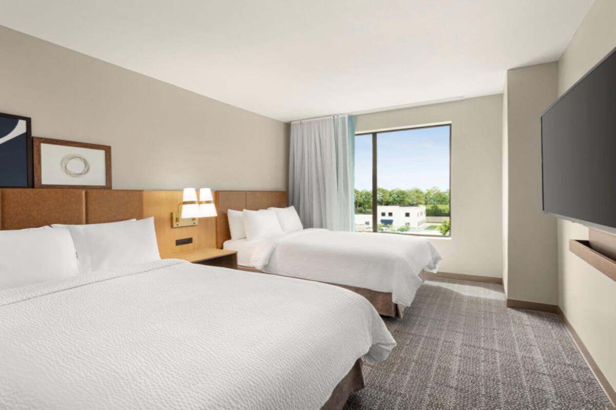 A hotel room with two beds, a large window, a flat-screen TV, and a framed picture on the wall.