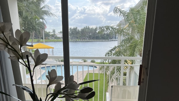 A balcony with a view of a body of water, a pool, and surrounding greenery.