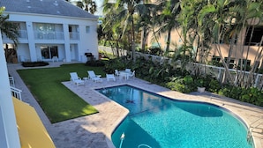 A swimming pool with a tiled deck, surrounded by a white fence, and adjacent to a building with a balcony.