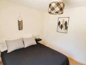 Un dormitorio con cama, almohadas, una mesita de noche con lámpara, un adorno en la pared y una obra de arte enmarcada.