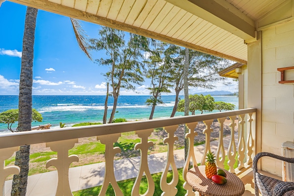 A balcony with a view of the ocean, palm trees, and a wicker chair with fruits. - Islander on the Beach 351 Direct Oceanfront Studio (Kapaa)