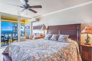 Quarto com uma cama grande, ventilador de teto, mesa de cabeceira com abajur e vista para o oceano através de uma porta de correr.