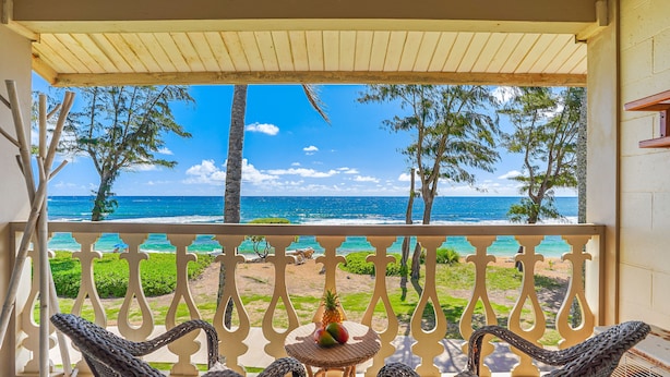 A balcony with wicker chairs, a small table with fruit, and a view of the ocean and beach. - View Beachfront, Split AC, Pool Hot Tub, King Bed (Kapaa)