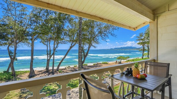 Un balcon avec vue sur l’océan et la plage, une table avec des fruits et une chaise.