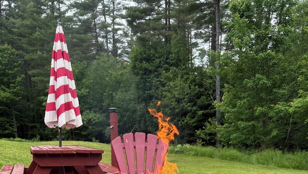 Una chimenea de piedra con una silla Adirondack roja y una mesa de picnic con un paraguas a rayas en un área exterior con césped.