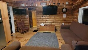 Interior de una cabaña de troncos con sofá, televisor montado en la pared y ventilador de techo.