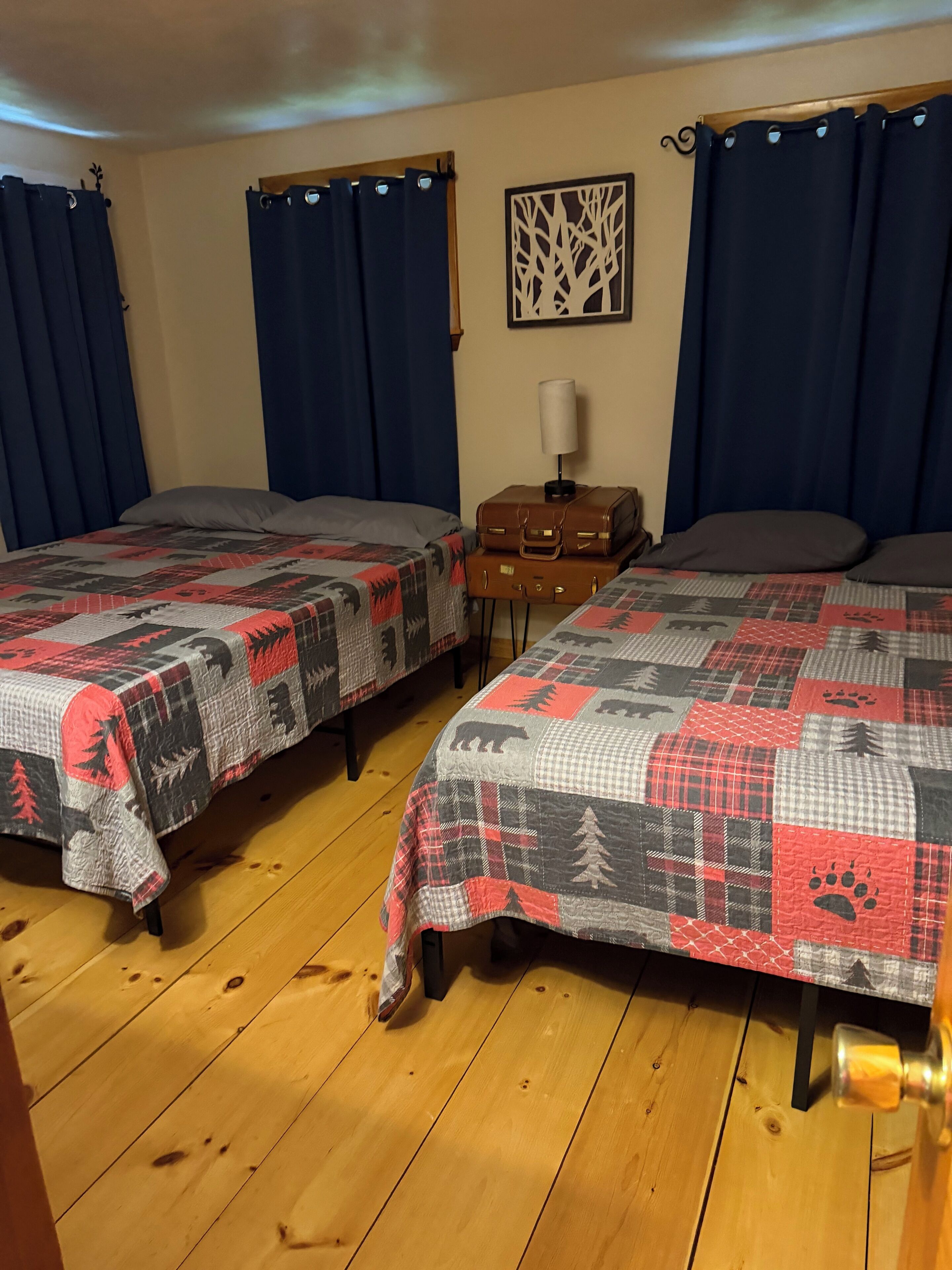 Quarto com duas camas, cada uma com colcha acolchoada estampada com padrões de animais. Há uma pequena mesa com abajur e uma mala entre as camas.