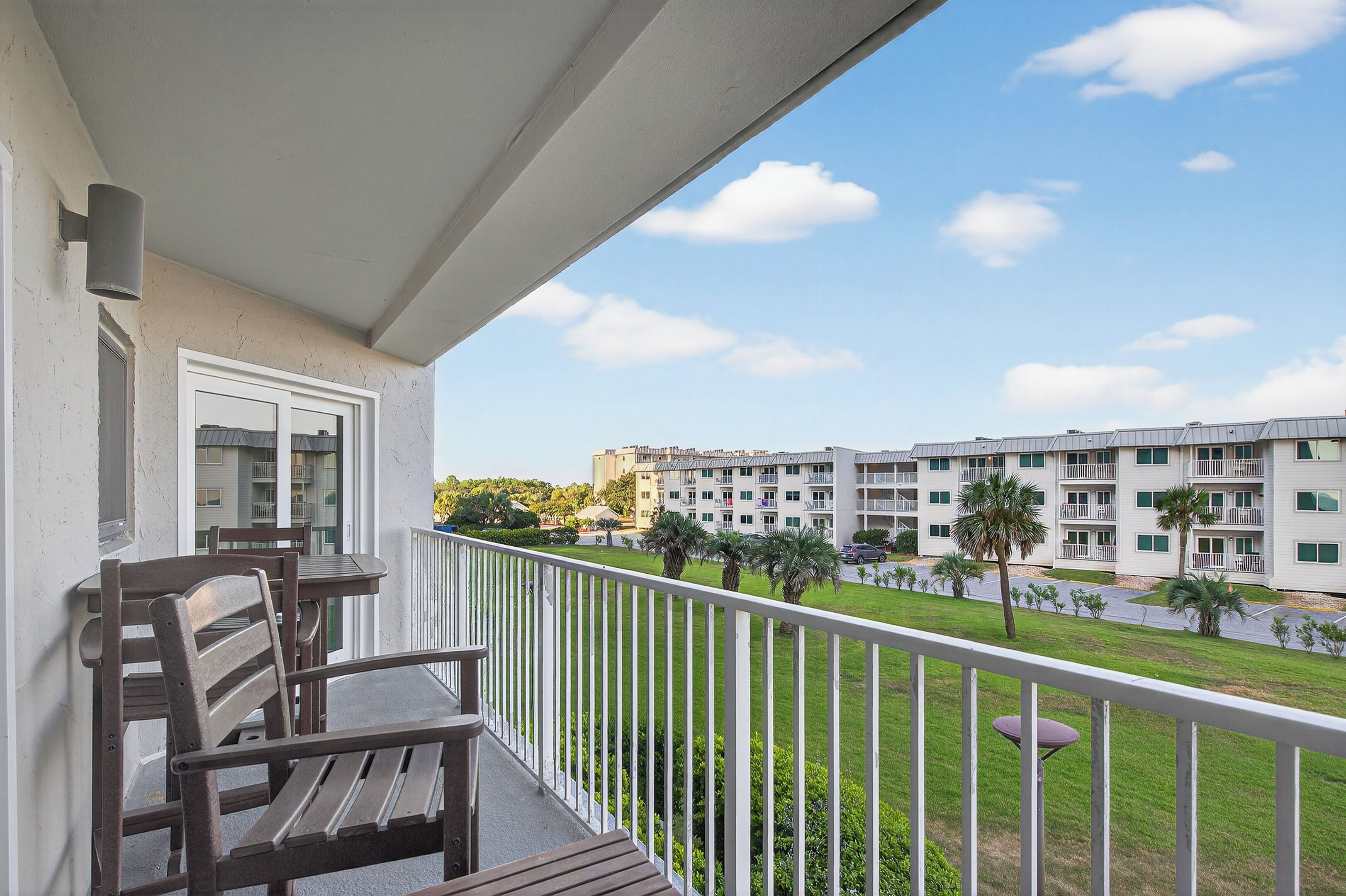 A balcony with a wooden table and chairs, overlooking a well-maintained lawn and a row of multi-story buildings with balconies.