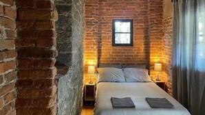 Ein Schlafzimmer mit Backsteinwand, Holzbalkendecke, einem Bett mit zwei Kissen, zwei Nachttischen mit Lampen und einem Fenster.