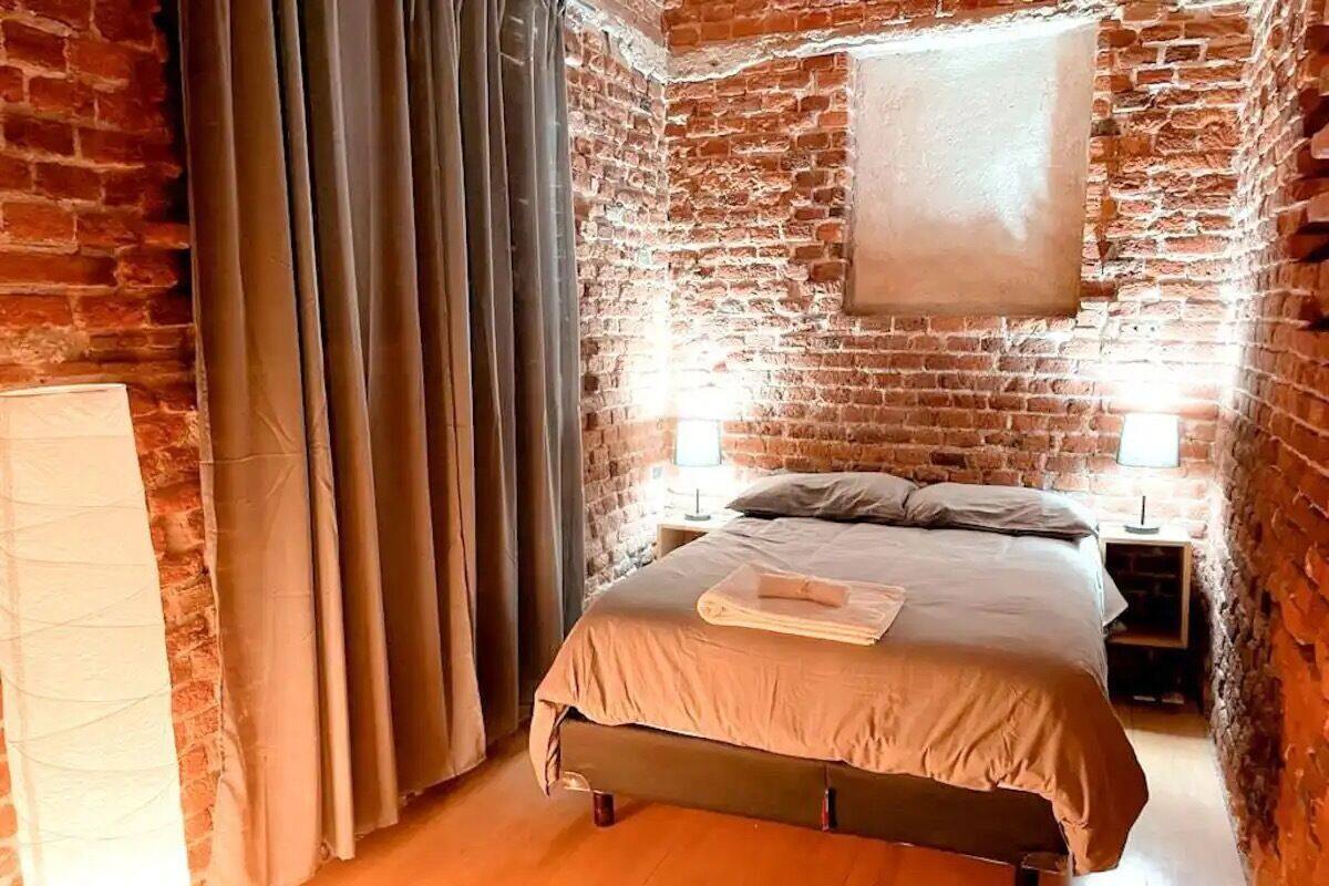 Un dormitorio con pared de ladrillo, una cama con colcha color beige, dos mesitas de noche con lámparas y una ventana con cortinas.
