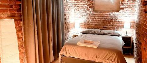 A bedroom with a brick wall, a bed with a beige bedspread, two bedside tables with lamps, and a window with curtains.