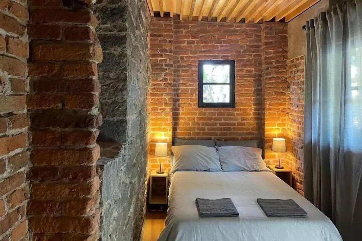 Una habitación con pared de ladrillo, techo de madera, cama con almohadas, dos mesitas de noche con lámparas y una ventana.