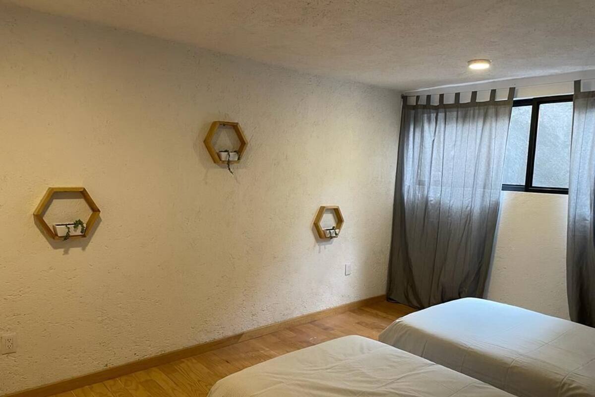 Una habitación con dos camas, suelo de madera, apliques de pared hexagonales, una ventana con cortinas e iluminación en el techo.