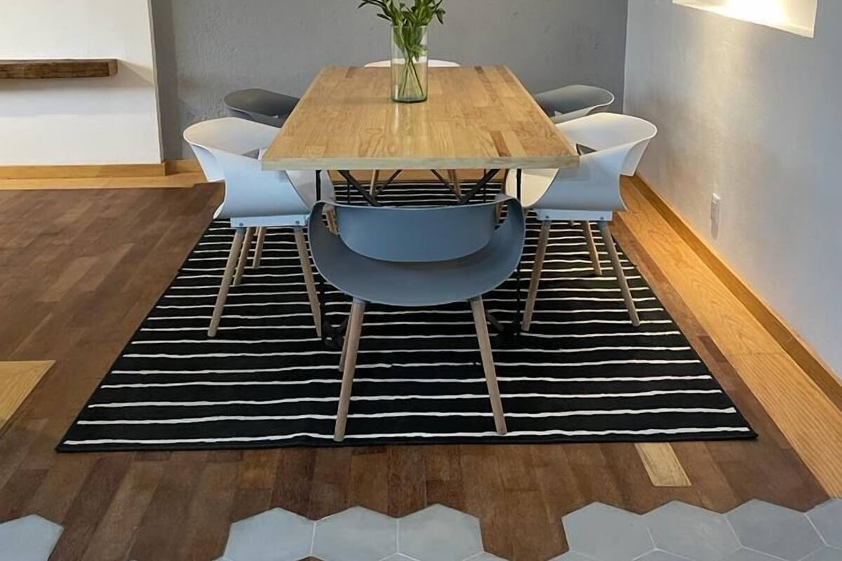 Un comedor con una mesa de madera, sillas, un florero con flores, un reloj en la pared y una alfombra con estampado geométrico.