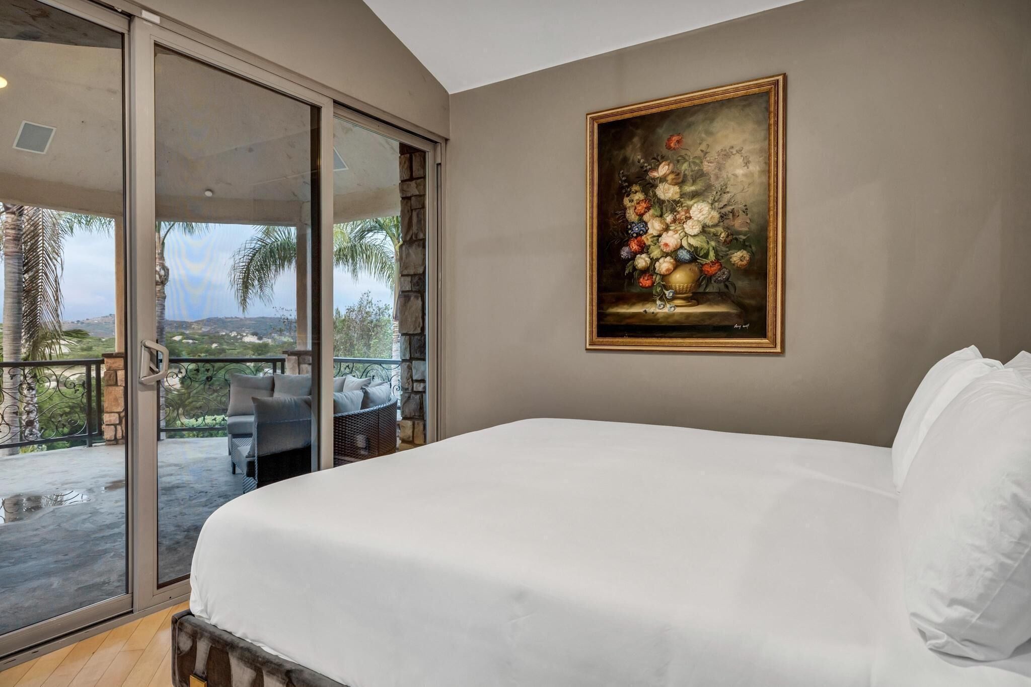 Une chambre avec un grand lit, un tableau représentant des fleurs et un balcon agrémenté de mobilier d’extérieur.