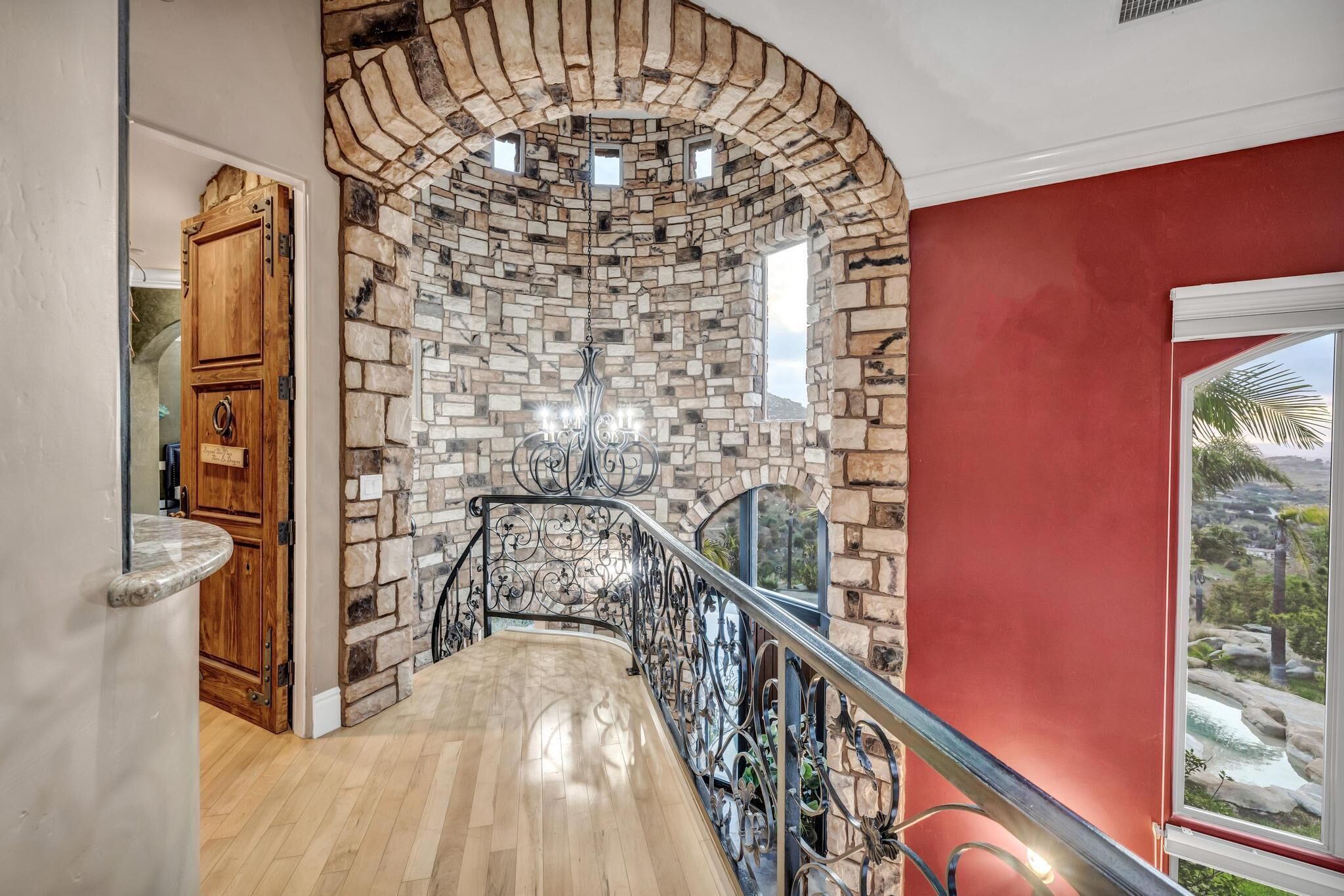 Una escalera con pared de piedra y barandilla de metal, que conduce a una habitación con pared roja y ventana con vistas al paisaje.