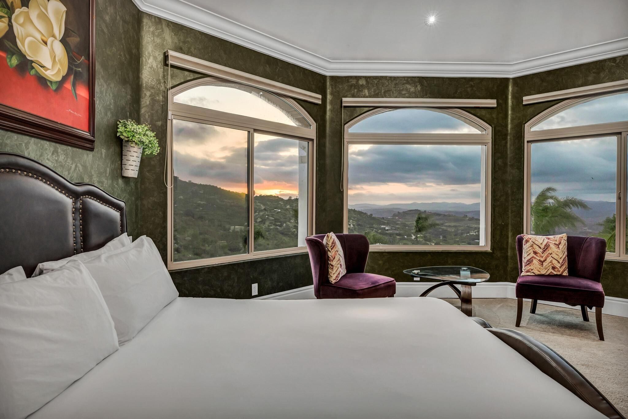 A bedroom with a large bed, two armchairs, a small table, and large windows offering a view of the landscape.