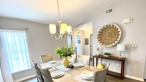 Uma sala de jantar com uma mesa de madeira posta para quatro pessoas, rodeada por cadeiras, um vaso de flores e um lustre.