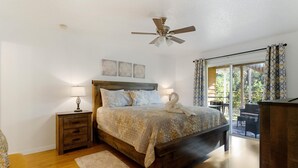 A bedroom with a wooden bed, two bedside tables with lamps, a ceiling fan, and a view of the outdoors through a sliding glass door.