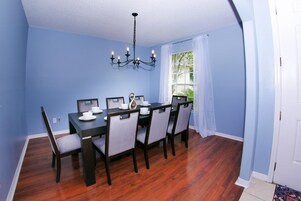A dining room with a dark wooden table and matching chairs, a chandelier, and a window with white curtains.
