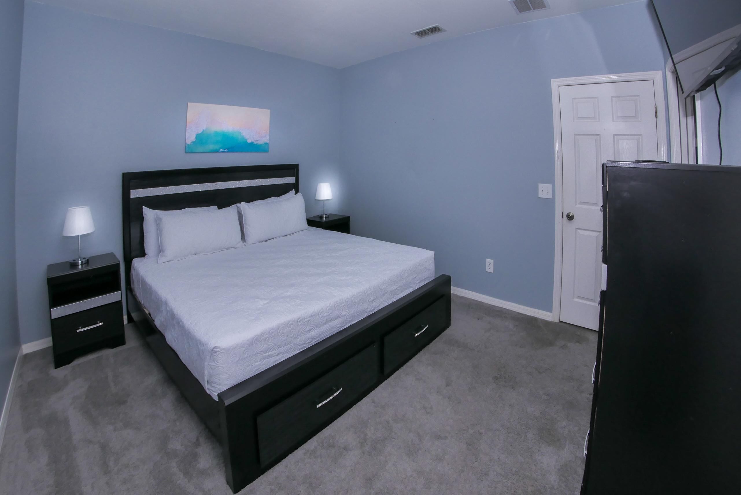 A bedroom with a bed, two bedside tables, a nightstand, a door, and a wall-mounted artwork.