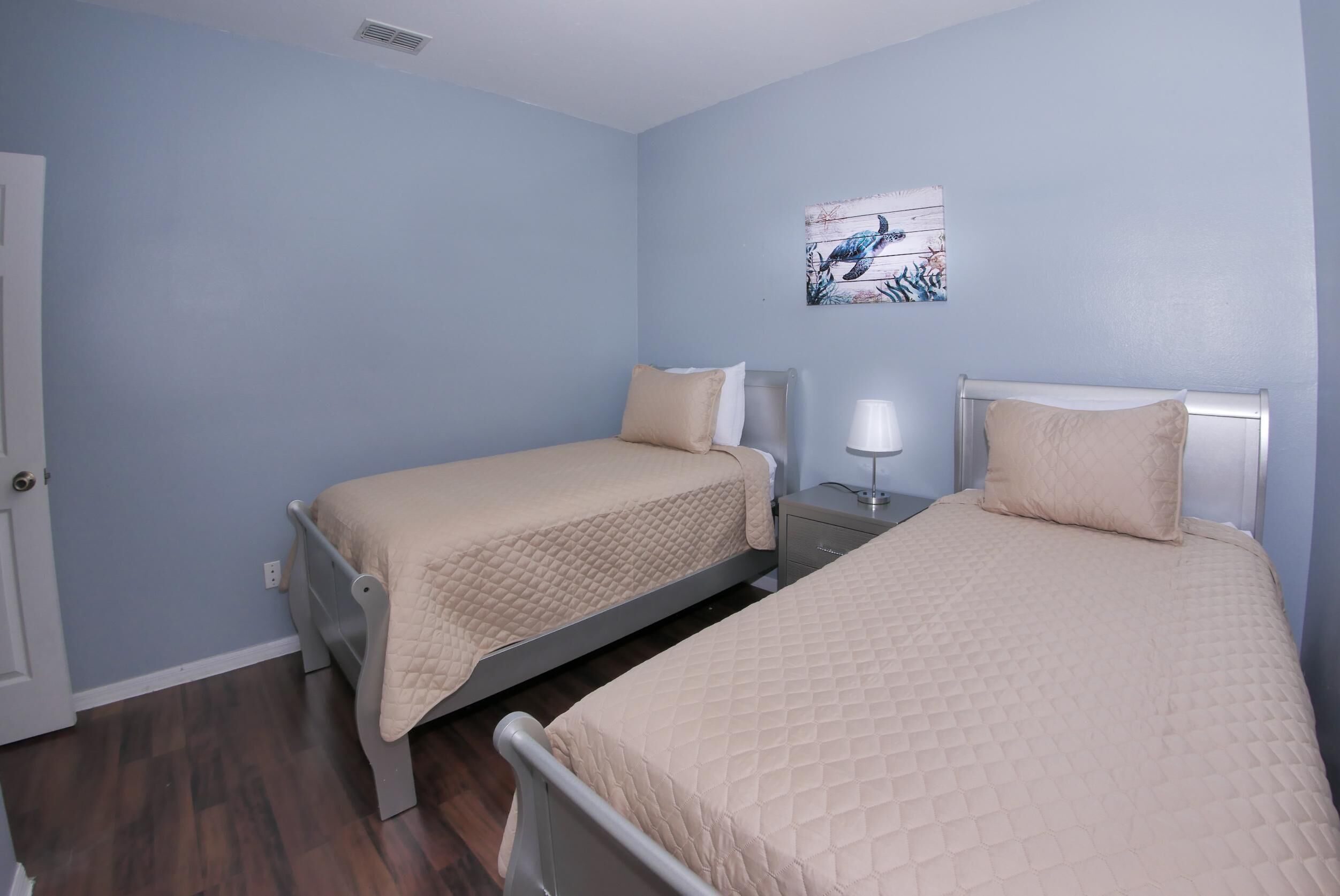 A bedroom with two beds, a nightstand, a wall-mounted lamp, and a door.