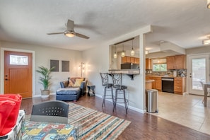 A spacious living area with a sofa, a coffee table, and a bar area with stools. There is a ceiling fan, a fireplace, and a kitchen with wooden cabinets and a refrigerator. - Topo House II: Fenced Yard & Fire Pit Oasis (Fruita)