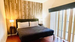 Une chambre à coucher avec une tête de lit en bois, un lit recouvert d’une couverture sombre, deux tables de chevet avec des lampes et une grande fenêtre avec des stores.