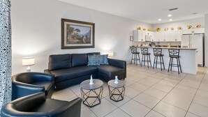 Un salon moderne avec un canapé noir, deux tables rondes et un coin cuisine doté de meubles blancs et de tabourets de bar.
