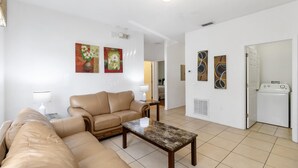 Un salon avec un canapé beige, une table basse et des œuvres d’art accrochées aux murs. Une porte donne accès à une buanderie.