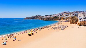 A beach with rows of sunbeds and umbrellas, a clear blue sea, and buildings on a hillside in the background.