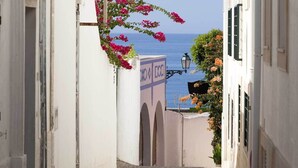 A narrow alleyway with white buildings, a sign with "MEO" and "ECU" on it, and a view of the sea.