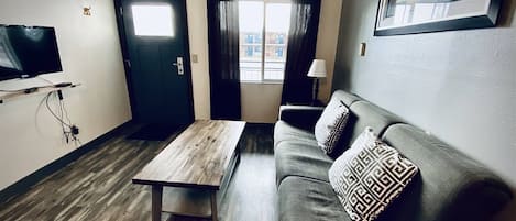A living room with a wooden coffee table, a sofa with patterned cushions, a television mounted on the wall, and a door leading to another room.