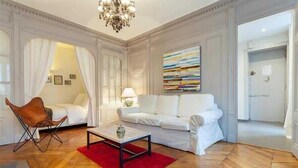 A room with a white sofa, a red rug, a coffee table, and a chandelier. - Colombe - Furnished apartment - Downtown Lyon (Lyon)