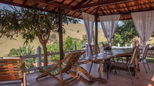 A covered outdoor seating area with a wooden table and chairs, surrounded by a white fence and trees. - Pousada Sonho Meu (Itanhandu)