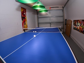 A blue ping pong table with a net, two white balls, and a green hanging lamp.