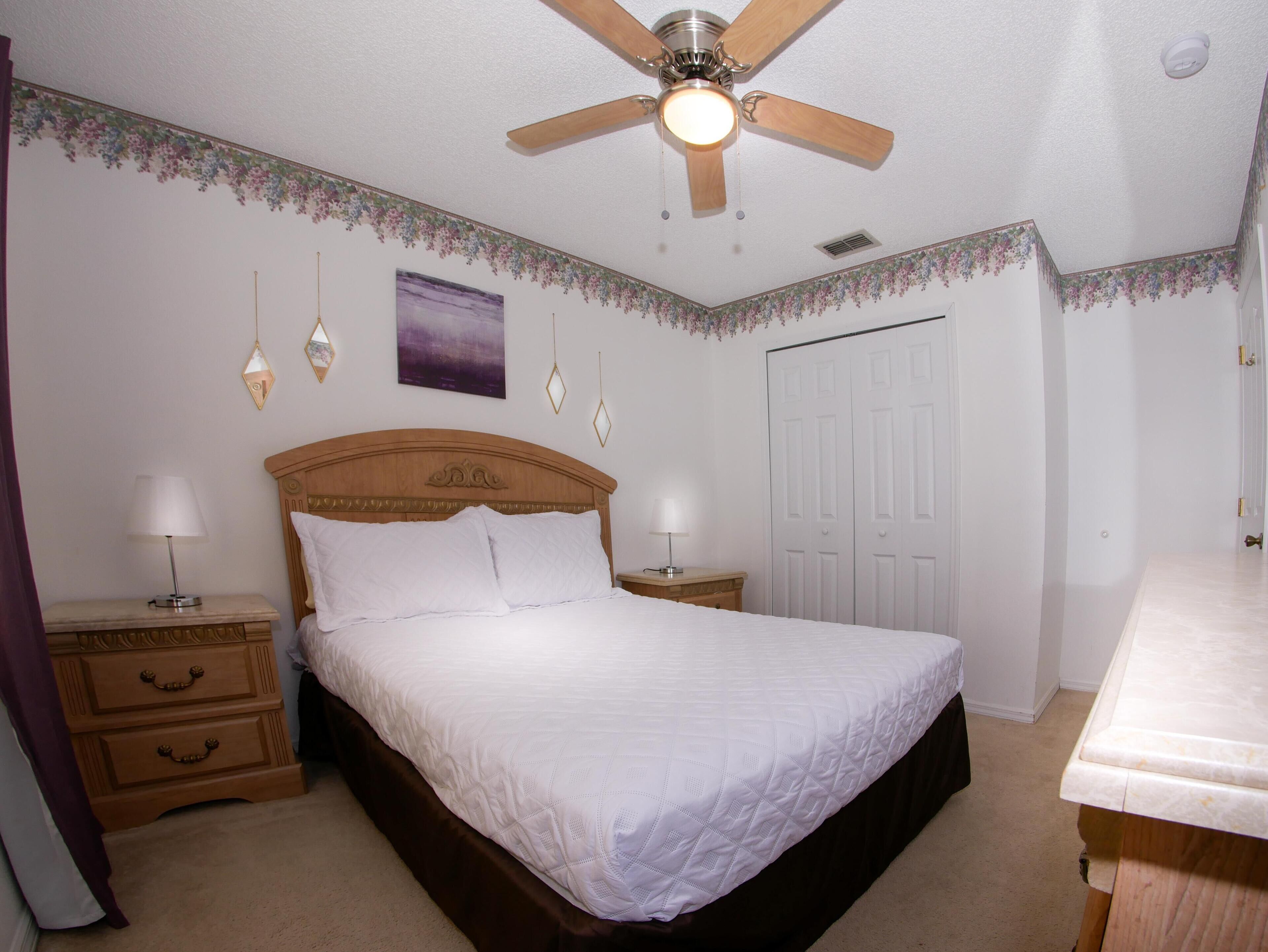 A bedroom with a wooden bed, two bedside tables with lamps, a ceiling fan, a door, and a wall-mounted light fixture.