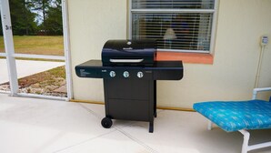 Een buitenbarbecue met drie bedieningsknoppen, een raam met luiken en een blauwe, met kussens beklede bank.