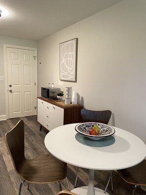 Een eethoek met een ronde tafel, twee stoelen, een magnetron, een kast en een abstract kunstwerk in een lijst aan de muur.