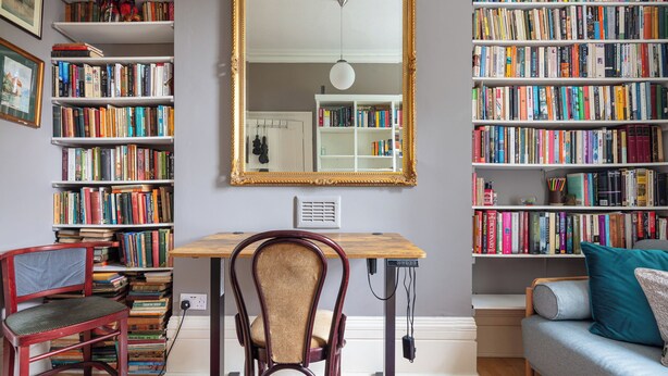 A study room with a wooden desk, a red chair, a bookshelf, a large mirror, and a grey sofa.