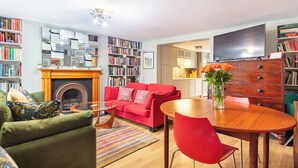 A living room with a fireplace, a red sofa, a coffee table, and a TV. There are bookshelves and a dining area with a wooden table and chairs.