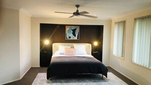 A modern bedroom with a bed, a ceiling fan, a painting on the wall, and a window with blinds. - Large Holiday Home with Heated Pool on demand near Lake & Beaches (Lake Munmorah)
