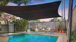 A rectangular swimming pool with a shaded area overhead, surrounded by a wooden deck and a metal fence. - Large Holiday Home with Heated Pool on demand near Lake & Beaches (Lake Munmorah)