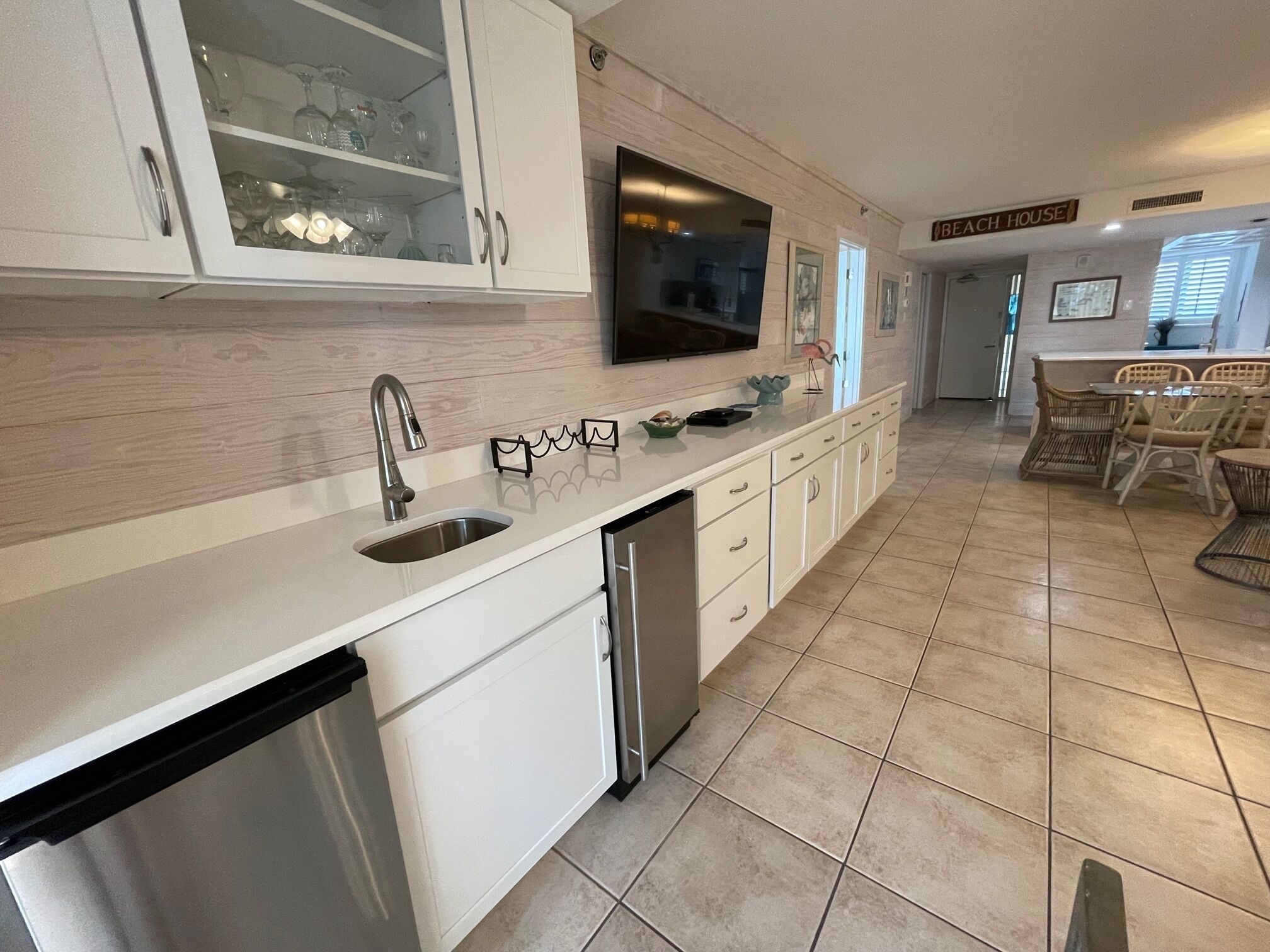 A modern kitchen with white cabinetry, a stainless steel sink, and a dishwasher. There is a dining area with wicker chairs and a table.