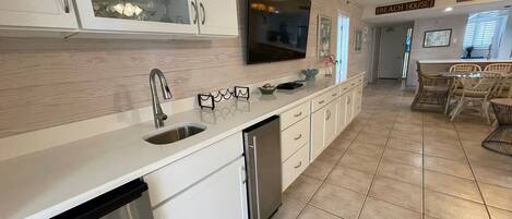A modern kitchen with white cabinetry, a stainless steel sink, and a dishwasher. There is a dining area with wicker chairs and a table.