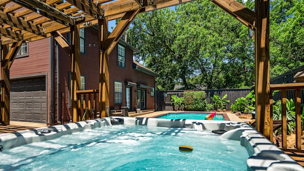 Un espace avec un jacuzzi, agrémenté d’une pergola en bois, une piscine et une maison en briques en arrière-plan.